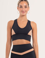 A woman modeling a black athletic sports bra with beige mesh side panels and a matching high-waisted black athletic skirt with a beige waistband, set against a plain light background.