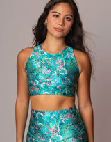 A woman modeling a matching two-piece activewear set featuring a vibrant turquoise blue floral print with pink, white, and green details. The set includes a halter crop top and high-waisted leggings, shown against a solid gray studio background.