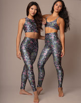 Two women modeling matching floral print activewear sets consisting of a sports bra and high-waisted leggings, standing against a solid beige studio background.
