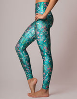 A model wearing high-waisted leggings with a shiny teal and green floral print, standing barefoot against a solid light beige background.