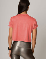 A woman shown from the back wearing a coral pink short-sleeve cropped t-shirt and metallic silver leggings, set against a plain light gray studio background.
