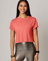 A woman wearing a coral pink short-sleeve crop top and metallic gray leggings, standing against a solid light gray studio background.