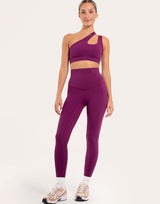 A woman modeling a matching athletic set consisting of a one-shoulder cutout sports bra and high-waisted leggings in a deep purple color, paired with white socks and multicolor sneakers, against a plain light background.