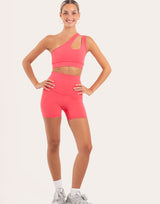 A woman modeling a bright pink activewear set consisting of a one-shoulder crop top with a side cutout and matching high-waisted biker shorts, paired with white socks and gray sneakers against a solid off-white background.
