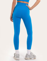 A model shown from the back wearing bright blue high-waisted leggings, a matching blue sports bra, white socks, and silver sneakers against a plain off-white studio background.