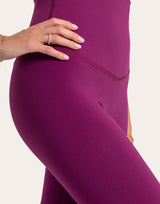Close-up of a model wearing high-waisted leggings in a rich plum purple color, showing the ribbed waistband and smooth fabric texture against a light off-white background.