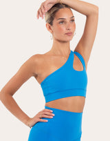 A woman modeling a bright blue one-shoulder crop top and matching high-waisted leggings against a light beige studio background.