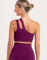 A woman shown from the back wearing a deep purple athletic sports bra and matching high-waisted leggings against a light off-white background.