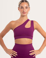 A woman modeling a matching athletic set featuring a one-shoulder crop top with a side cutout and high-waisted leggings, in a solid deep purple color, against a light beige studio background.
