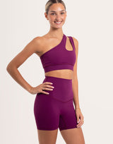 A woman modeling a matching athletic set consisting of a one-shoulder crop top and high-waisted biker shorts in a deep purple color, set against a plain light background.
