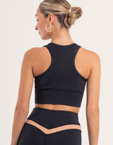 Rear view of a woman wearing a black athletic crop top and matching high-waisted leggings with a beige V-cut waistband detail, set against a plain light background.