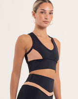 A woman modeling a black athletic sports bra with sheer mesh side panels and a matching high-waisted black activewear bottom against a light beige studio background.