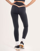 A model from the back wearing black high-waisted leggings with a peach-colored V-shaped waistband detail, paired with white socks and black sneakers with orange accents, against a plain off-white background.