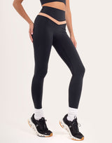 A model wearing black high-waisted leggings with a beige waistband accent, paired with white socks and black running shoes, shown from the waist down on a light studio background.