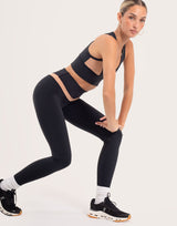 A woman in a black athletic crop top and matching black leggings poses in a lunge position against a clean, light off-white studio background. She is wearing black sneakers with white socks.