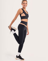 A woman modeling a black athletic set consisting of a sports bra with mesh side panels and matching high-waisted leggings, posing in a studio setting.