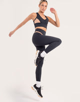 A woman in a black athletic crop top and matching high-waisted leggings jumps against a light gray studio background. She is wearing black sneakers with white and orange accents, white socks, and has gold hoop earrings.