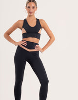 A woman modeling a black athletic set consisting of a sports bra with side cutouts and matching high-waisted leggings, posed against a solid off-white studio background.