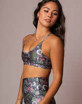 A woman modeling a matching two-piece activewear set featuring a metallic silver base with a vibrant pink, green, and blue floral print, set against a solid taupe background.