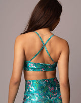 A woman shown from the back wearing a teal floral two-piece set with a matching crop top and skirt. The top features criss-crossing straps, and the floral pattern contains shades of pink, white, and green on a teal background.