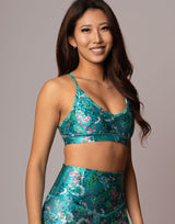 A woman modeling a matching teal floral sports bra and high-waisted leggings set against a solid gray studio background.