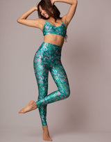 A woman modeling a matching teal and green floral print activewear set consisting of a sports bra and high-waisted leggings, posed against a solid light gray studio background.