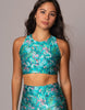 A woman modeling a matching two-piece activewear set featuring a vibrant turquoise blue floral print with pink, white, and green details. The set includes a halter crop top and high-waisted leggings, shown against a solid gray studio background.