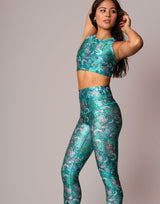 A woman modeling a matching teal green athletic set with a floral print, featuring a sleeveless crop top and high-waisted leggings, against a solid light gray background.