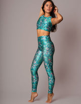 A woman modeling a matching two-piece activewear set with a teal green and pink floral print, consisting of a crop top and high-waisted leggings, against a solid gray studio background.
