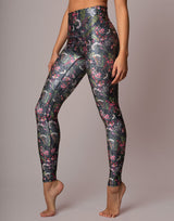 A model wearing high-waisted leggings with a dark green and black background featuring a detailed print of birds, flowers, and leaves, posed against a solid light gray studio background.