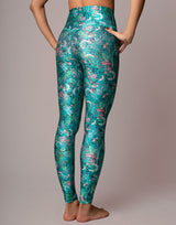 A model from the waist down wearing high-waisted leggings with a teal and green floral branch pattern, standing against a solid light gray studio background.