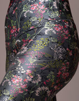 Close-up of a person wearing shiny black leggings with a dense floral print in pink, white, green, and yellow, against a plain taupe background.