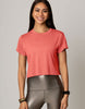 A woman wearing a coral pink short-sleeve crop top and metallic gray leggings, standing against a solid light gray studio background.