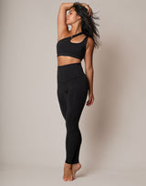 A woman modeling a matching black activewear set consisting of a one-shoulder cutout sports bra and high-waisted leggings, standing against a plain beige studio backdrop.