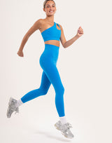 A woman running in a bright blue activewear set consisting of a one-shoulder sports bra and high-waisted leggings, paired with white socks and silver sneakers, against a plain off-white studio background.
