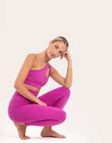 A woman in a bright magenta activewear set consisting of a sports bra and leggings, crouching in a yoga pose against a plain off-white studio background.