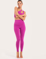 A woman modeling a bright magenta activewear set consisting of a one-shoulder sports bra and high-waisted leggings, standing against a plain light beige studio background.