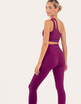 A woman modeling a matching athletic set consisting of a magenta-purple sports bra and high-waisted leggings, shown from a three-quarters back angle against a solid off-white background.