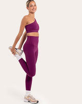 A woman in a matching deep purple activewear set, consisting of a one-shoulder sports bra and high-waisted leggings, performing a side stretch with one leg bent backward. She is wearing white socks and gold and white athletic sneakers against a solid off-white background.