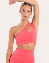 A woman modeling a bright coral pink athletic set, featuring a one-shoulder cutout sports bra and matching high-waisted leggings, against a solid light beige background.