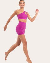 A woman modeling a bright pink activewear set consisting of a one-shoulder crop top and matching high-waisted shorts, posed against a solid off-white background.