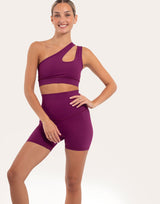 A woman modeling a matching athletic set consisting of a one-shoulder cutout sports bra and high-waisted biker shorts in a deep purple color, set against a plain light background.