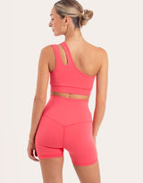 A woman shown from the back wearing a bright coral pink activewear set consisting of a one-shoulder sports bra and matching high-waisted biker shorts, against a light off-white studio background.