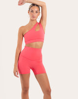A woman modeling a bright coral pink two-piece activewear set consisting of a one-shoulder cutout sports bra and matching high-waisted biker shorts, set against a solid off-white background.