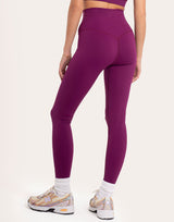 A model from the back wearing high-waisted, form-fitting athletic leggings in a deep plum purple color, paired with white socks and beige and white sneakers, against a solid off-white background.
