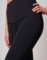 Close-up of a model wearing black high-waisted maternity leggings with a wide, supportive waistband and a side pocket, set against a neutral light gray background.