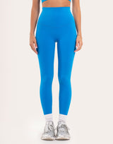 A model from the waist down wearing bright blue high-waisted leggings, white socks, and silver sneakers against a plain off-white studio background.
