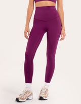 A model wearing high-waisted, form-fitting leggings in a deep plum purple color, paired with white socks and athletic sneakers, against a plain off-white studio background.