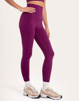 A model wearing high-waisted, form-fitting leggings in a deep plum purple color, paired with white socks and New Balance sneakers, shown from the waist down against a plain off-white studio background.