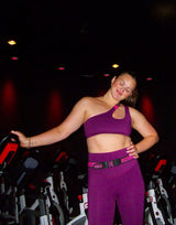 A woman in a purple activewear set consisting of a one-shoulder crop top and matching leggings, posing in a gym setting with exercise bikes in the background.